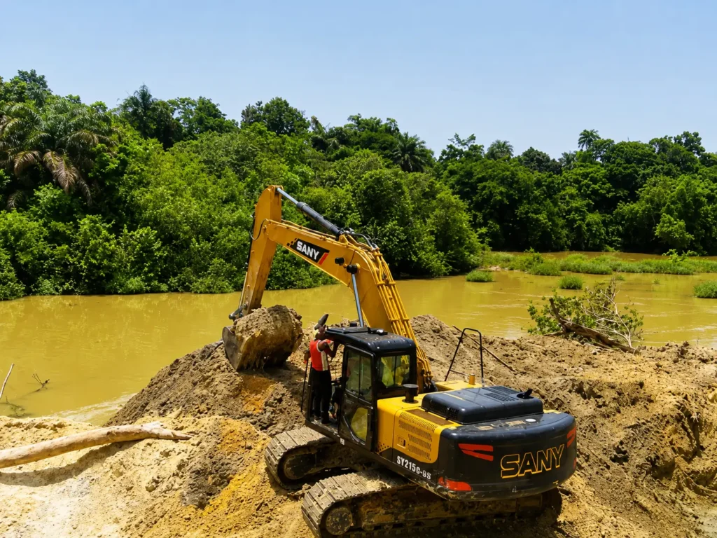 A yellow SANY hydraulic excavator digging up earth next to a turbid, muddy river in Rosinth village, Sierra Leone.