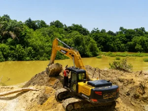 A yellow SANY hydraulic excavator digging up earth next to a turbid, muddy river in Rosinth village, Sierra Leone.